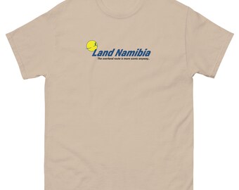 May include: A tan short-sleeve t-shirt with the text "Land Namibia" in blue and a yellow graphic. Below the text is the phrase "The overland route is more scenic anyway." The shirt is made of a soft material.