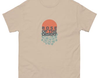May include: A tan t-shirt with a graphic design. The design features a red-orange circle above the words "ROSE OF THE DESERT" in gray and teal. Below the text is a teal and tan floral design.