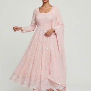 May include: A light pink Anarkali suit with intricate floral embroidery and sequin embellishments. The long-sleeved dress features a square neckline and a flowing skirt, paired with a matching dupatta and pants. The outfit is completed with embellished flats.