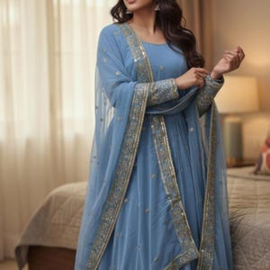 May include: A light blue dress with a matching sheer shawl. The dress features gold and silver embroidered trim along the sleeves and edges of the shawl. The dress has a flowing, pleated design.