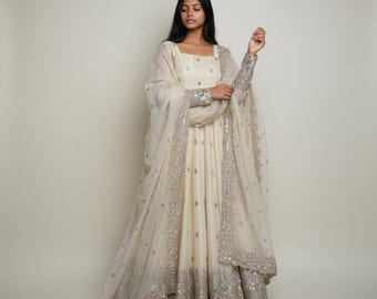 Op de afbeelding: Een crèmekleurige Anarkali-jurk die tot op de grond valt, voorzien van ingewikkeld zilverkleurig borduurwerk langs de zoom, manchetten en halslijn. De outfit bevat een bijpassende transparante dupatta die over de schouders is gedrapeerd.
