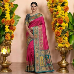 May include: A woman wearing a vibrant pink sari with intricate gold and teal border detailing. The sari features a detailed pattern along the border and a contrasting gray blouse. The background includes floral arrangements and a decorative lamp.