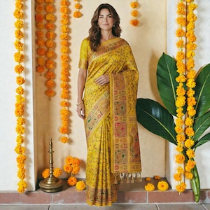 May include: A woman in a yellow saree with a detailed floral pattern and a decorative border. The saree is draped over the body. The background features marigold garlands and a brass lamp.