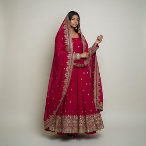 Peut inclure: Une robe Anarkali rouge vif avec des broderies dorées complexes. L'ensemble comprend une dupatta assortie drapée sur la tête et les épaules, également ornée de détails dorés. La robe présente une silhouette fluide et des ornements.