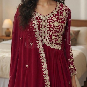 Puede incluir: Vestido color burdeos con intrincados bordados dorados en el corpiño y las mangas. El vestido presenta un escote en V y una falda fluida. El bordado incluye patrones florales y de hojas, y tiene un chal transparente a juego.