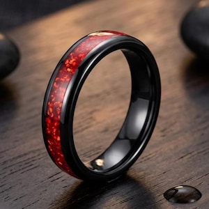 Puede incluir: Un anillo negro con una banda lisa y pulida que presenta una incrustación central de material rojo vibrante y triturado. El anillo tiene un perfil redondeado y un acabado brillante, mostrando un contraste llamativo entre el metal oscuro y la incrustación roja brillante.