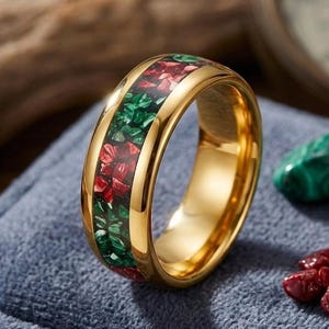 Könnte beinhalten: Ein goldfarbener Ring mit einer zentralen Einlage aus zerkleinerten roten und grünen Steinfragmenten. Das Band hat eine polierte, reflektierende metallische Oberfläche, und die Einlage besteht aus kleinen, unregelmäßigen Stücken aus rotem und grünem Material, die in einem durchgehenden Muster um den gesamten Umfang des Rings angeordnet sind.