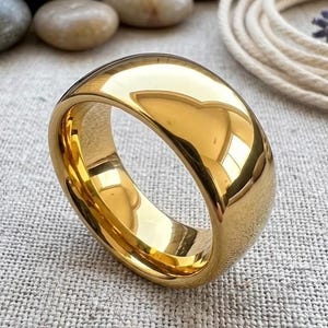 May include: A polished gold ring with a rounded, smooth design. The ring reflects light, giving it a bright appearance. The ring is a simple band, suitable for everyday wear. The ring is a jewelry item.