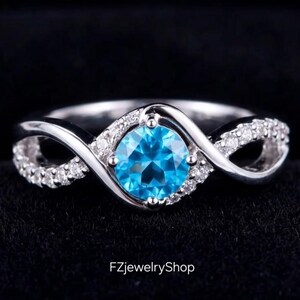 Pode incluir: Um anel de prata com uma pedra preciosa redonda azul brilhante no centro. A banda apresenta um design infinito com pequenas pedras transparentes ao longo das curvas. O anel é exibido em uma superfície preta, com o texto "FZjewelryShop".