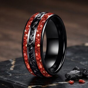 Puede incluir: Un anillo negro con una incrustación central de fragmentos metálicos oscuros y texturizados, flanqueado por dos bandas de material rojo vibrante triturado. El anillo tiene un acabado pulido y liso en los bordes exteriores.