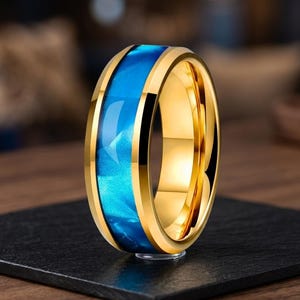 May include: A gold-toned ring featuring a polished metallic band with a central inlay of vibrant blue, swirling resin. The ring has a smooth, reflective finish on the outer edges and a bright, glossy appearance throughout the entire circular band.