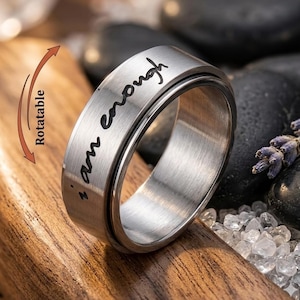 Peut inclure: Une bague rotative en métal argenté avec une finition brossée. La bande extérieure présente le texte noir gravé "i am enough" et un graphique en forme de flèche incurvée avec le mot "Rotatable" indiquant le mouvement rotatif de la bague.
