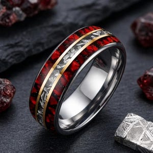 May include: A metallic ring featuring a central band of silver-toned textured material flanked by two thin gold-toned lines. The outer edges of the ring are inlaid with a deep red, fragmented pattern, all set within a polished silver-toned metal band.