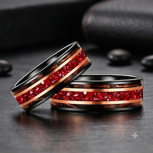 Puede incluir: Dos anillos con banda negra, una incrustación central de piedras rojas trituradas y finos detalles metálicos en color cobre. El diseño incluye una textura de veta de madera oscura que rodea la incrustación de piedras rojas.