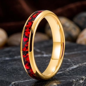 May include: A gold-toned ring featuring a central inlay of dark material embedded with small, irregular bright red fragments. The band has a smooth, polished metallic finish and a rounded profile, showcasing a distinct contrast between the gold metal and the red inlay.