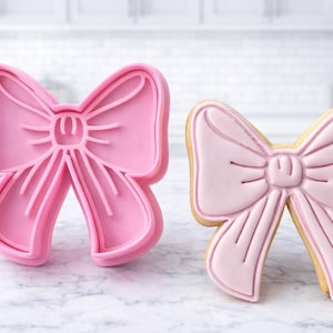 Op de afbeelding: Een roze strikvormige koekjesuitsteker naast een versierd strikvormig koekje. Het koekje is versierd met roze glazuur en een lichtbruine rand. De achtergrond is een witte keuken.