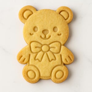 Può includere: Un biscotto a forma di orsetto color marrone dorato con un papillon. Il biscotto presenta dettagli in rilievo per gli occhi, il naso, la bocca e il papillon. Le braccia e le gambe dell'orso sono arrotondate. Il biscotto è su una superficie bianca.