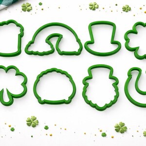Può includere: Otto formine verdi in plastica per biscotti a forma di simboli di San Patrizio, tra cui un boccale di birra, un arcobaleno, un cappello a cilindro, un quadrifoglio e un ferro di cavallo. Le formine sono su una superficie bianca con coriandoli.