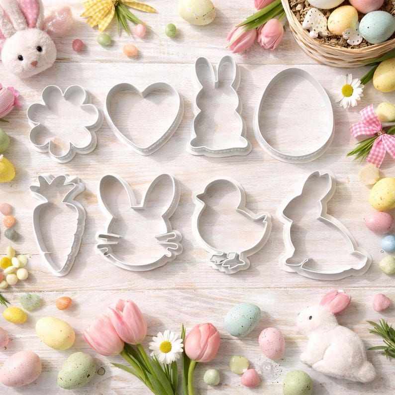 May include: A collection of white metal cookie cutters in Easter shapes, including a flower, heart, bunny, egg, carrot, bunny face, chick, and another bunny. The cutters are arranged on a light wood surface with Easter decorations.