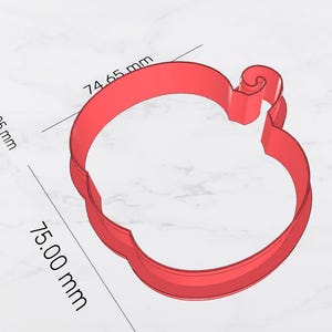 May include: A red apple-shaped cookie cutter. The cutter is approximately 3 inches wide and 3 inches tall. The area is 3,905 mm. The apple has a stem and a curved top. The cutter is made of a solid material.