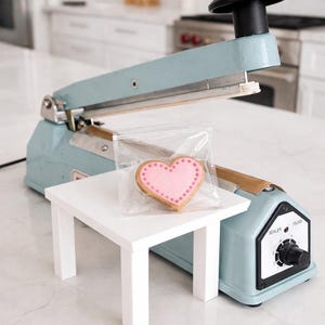 May include: A light blue heat sealer with a black handle and a white table holding a heart-shaped cookie in a clear bag. The cookie has a pink center and a brown border with pink dots.