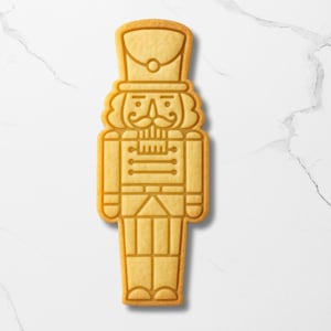 May include: A golden-brown cookie in the shape of a nutcracker soldier. The cookie has detailed line work depicting the soldier's uniform, hat, and facial features, including a mustache. The cookie is set against a white marble background.