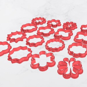 May include: A collection of red plastic cookie cutters in various floral and decorative shapes. The cutters are arranged on a white surface, with some text visible on the sides. The shapes include flowers, scalloped ovals, and other stylized designs.