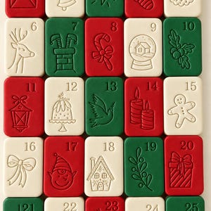 May include: A rectangular advent calendar set featuring twenty-five individual tiles in red, green, and white. Each tile is embossed with a unique holiday-themed icon and a sequential number from one to twenty-five, including images of a letter to Santa, a bell, a star, a stocking, a wreath, a reindeer, a chimney, a candy cane, a snow globe, holly, a lantern, a cake, a dove, candles, a gingerbread man, a bow, an elf, a house, a branch, a gift, an ornament, a sleigh, a candy, Santa, and a tree.