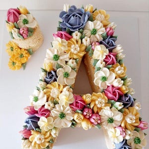 May include: A cake shaped like the letter "A", lavishly decorated with buttercream flowers in various colours, including yellow, pink, white, and blue. A smaller, matching floral arrangement is beside it. The cake is on a white surface.