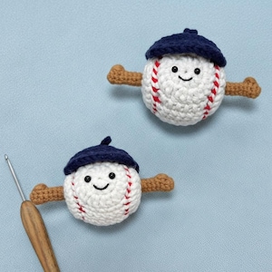 May include: Two crocheted baseballs with smiling faces, red stitching, and navy blue caps. Each baseball has brown arms extending from the sides. The baseballs are on a light blue background.