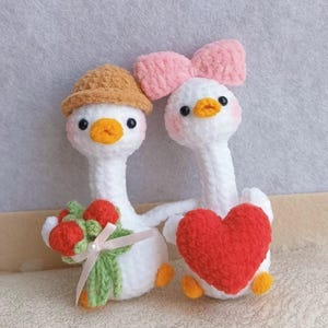 以下が含まれることがあります： 手作りの編み物のアヒル2羽。1羽は茶色の帽子と赤い花のブーケ、もう1羽はピンクのリボンと赤いハートを持っています。アヒルは白い体、オレンジ色のくちばし、黒い目です。魅力的で気まぐれなデザインです。