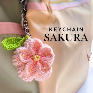 Puede incluir: Un llavero de ganchillo con una flor de sakura rosa con centro amarillo y una hoja verde. El llavero está sujeto a una cadena plateada. El texto "KEYCHAIN SAKURA" se muestra en negro.