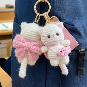 May include: Two white crochet cat keychains with pink accents. One cat has a pink bow, the other a pink purse. The keychains are attached to a gold-colored ring, hanging against a blue backpack.