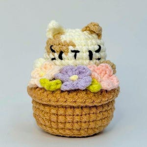 May include: A crocheted cat figurine with a basket of flowers. The cat has white and brown fur, closed eyes, and the word "Sweet" stitched on its face. The basket is brown, and the flowers are pink, purple, and yellow.