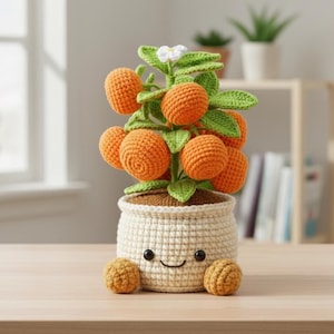 Puede incluir: Un árbol de naranjos tejido a ganchillo en una maceta sonriente. El árbol presenta naranjas tejidas a ganchillo de color naranja brillante, hojas verdes y una pequeña flor blanca. La maceta es de color blanquecino con una cara sonriente y dos bolitas naranjas.