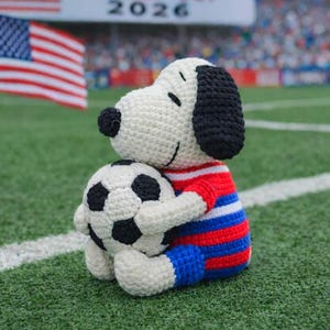 May include: A crocheted Snoopy doll wearing a red, white, and blue outfit, hugging a soccer ball. The background includes an American flag and a banner that reads "WORLD CUP 2026". The doll is on a green field.