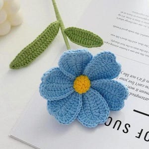 May include: A handmade crochet flower with a light blue petal design and a yellow center. The flower is attached to a green stem with green leaves. The flower is resting on a white surface with text.
