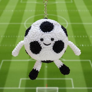 May include: A handmade, crocheted soccer ball keychain with a smiling face, black and white design, and attached metal keyring. The soccer ball has arms and legs, giving it a playful appearance. The background is a green soccer field.