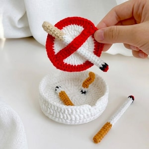 Puede incluir: Un cenicero de ganchillo blanco que contiene dos pequeñas colillas de cigarrillo de ganchillo, acompañado de un cigarrillo de ganchillo blanco separado y un letrero circular de prohibido fumar en rojo y blanco con un cigarrillo fijado.