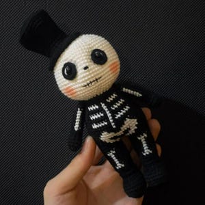 Op de afbeelding: Een handgemaakte gehaakte skeletpop met een zwarte hoge hoed. De pop heeft een witte kop met zwarte ogen en een gestikte mond. Het lichaam van het skelet is zwart met witte botdetails. De wangen van de pop zijn roze.
