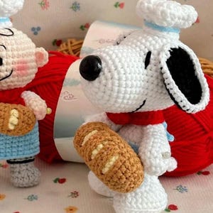 May include: Crocheted Snoopy figure wearing a chef's hat and red scarf, holding a loaf of bread. The figure is white with black ears and nose. Another crocheted figure is visible on the left, wearing a chef's hat and holding a loaf of bread.