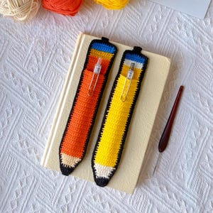 May include: Two crochet pencil-shaped pen cases, one orange and one yellow, each featuring a black border, a blue band at the top, and a beige tip. Each case includes a silver metal pen clip attached to the top section, resting on a closed book.