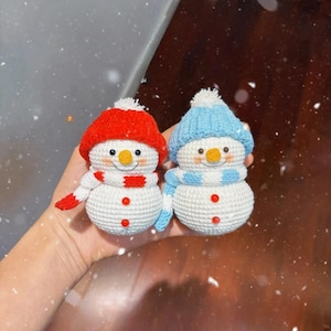 May include: Two handmade crochet snowmen. One snowman wears a red hat and scarf, the other a blue hat and scarf. Both have orange noses, black eyes, and red button details. The snowmen are white with a textured appearance.