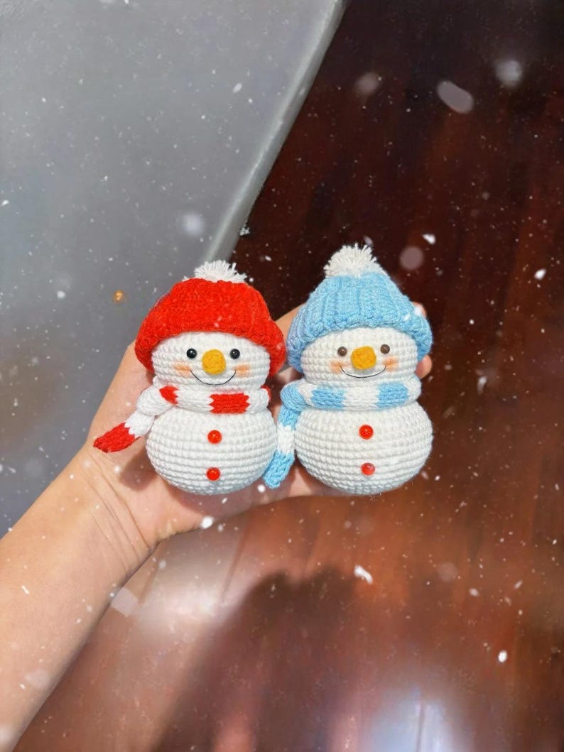 May include: Two handmade crochet snowmen. One snowman wears a red hat and scarf, the other a blue hat and scarf. Both have orange noses, black eyes, and red button details. The snowmen are white with a textured appearance.