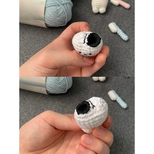 K&ouml;nnte beinhalten: Nahaufnahme eines wei&szlig;en geh&auml;kelten Auges mit schwarzer Pupille, gehalten in einer Hand. Das Auge ist Teil eines gr&ouml;&szlig;eren H&auml;kelprojekts, mit blauer Wolle und anderen H&auml;kelteilen im Hintergrund.