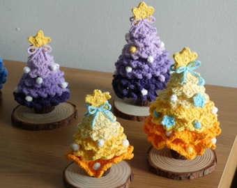 Op de afbeelding: Gehaakte kerstbomen in paars, geel en oranje, elk versierd met een gele ster en witte of blauwe accenten. De bomen staan op kleine houten schijfjes, wat een feestelijke en handgemaakte uitstraling geeft. Elke boom is ongeveer 10 cm hoog.