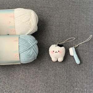 K&ouml;nnte beinhalten: Ein H&auml;kel-Zahn- und Zahnb&uuml;rsten-Charm-Set mit wei&szlig;er und hellblauer Wolle. Der Zahn-Charm ist wei&szlig; mit einem schwarzen Fleck und rosa Wangen. Die Zahnb&uuml;rste ist hellblau. Die Wolle ist wei&szlig; und hellblau.