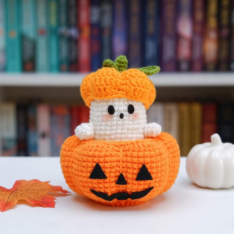 May include: A crocheted ghost figure wearing an orange pumpkin hat, sitting inside a larger orange crocheted jack-o'-lantern with black felt eyes and a mouth. The ghost has a white body, two small black eyes, and a tiny black stitched mouth.