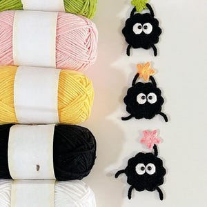 Puede incluir: Una colecci&oacute;n de ovillos de hilo de colores en verde, rosa, amarillo, negro y blanco. Junto al hilo, tres criaturas negras de ganchillo con ojos blancos y detalles en forma de estrella, ideales para proyectos de manualidades.