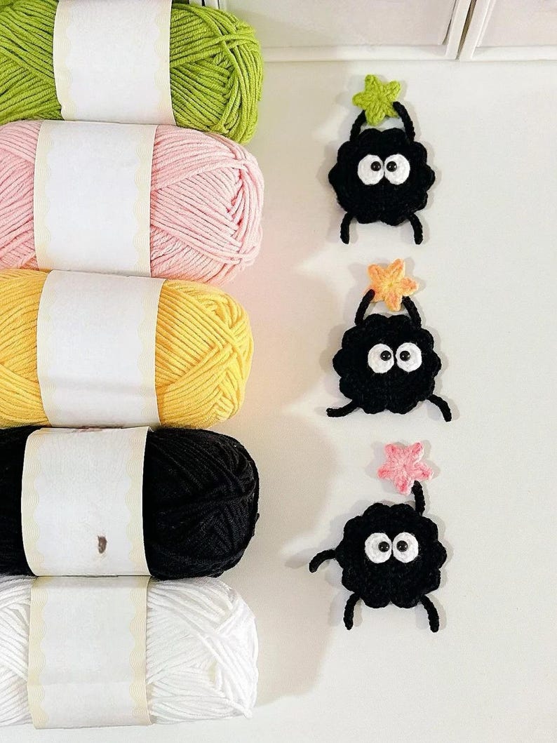Puede incluir: Una colecci&oacute;n de ovillos de hilo de colores en verde, rosa, amarillo, negro y blanco. Junto al hilo, tres criaturas negras de ganchillo con ojos blancos y detalles en forma de estrella, ideales para proyectos de manualidades.